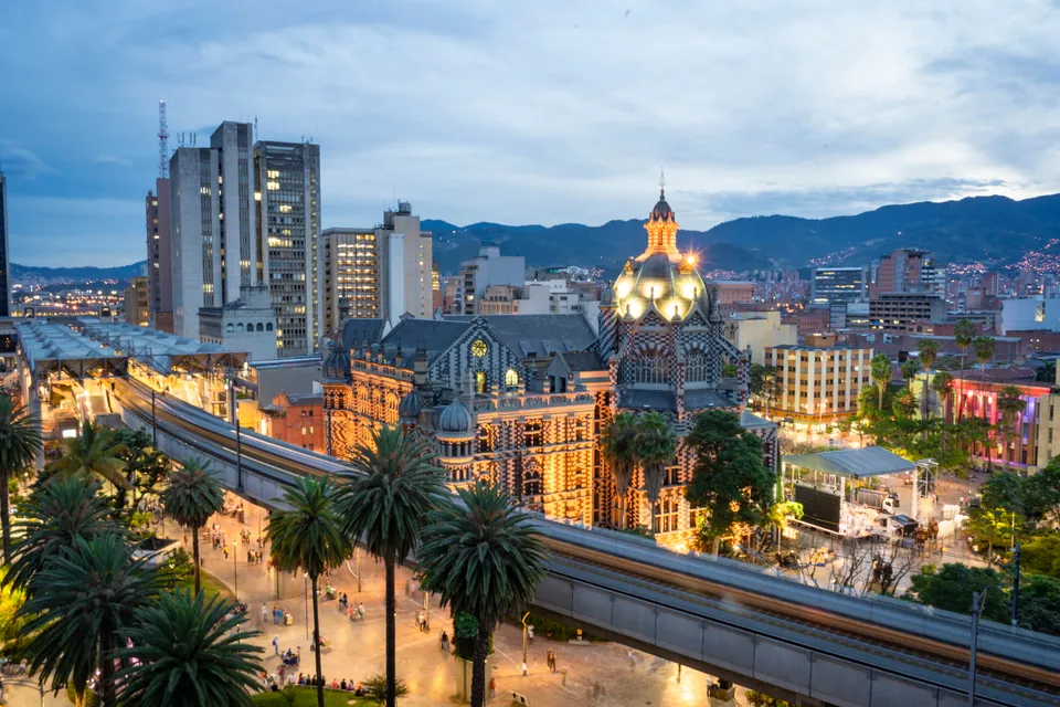 Centro de Medellín al anochecer: el Palacio de la Cultura iluminado, la línea del Metro entre palmeras y las montañas al fondo.