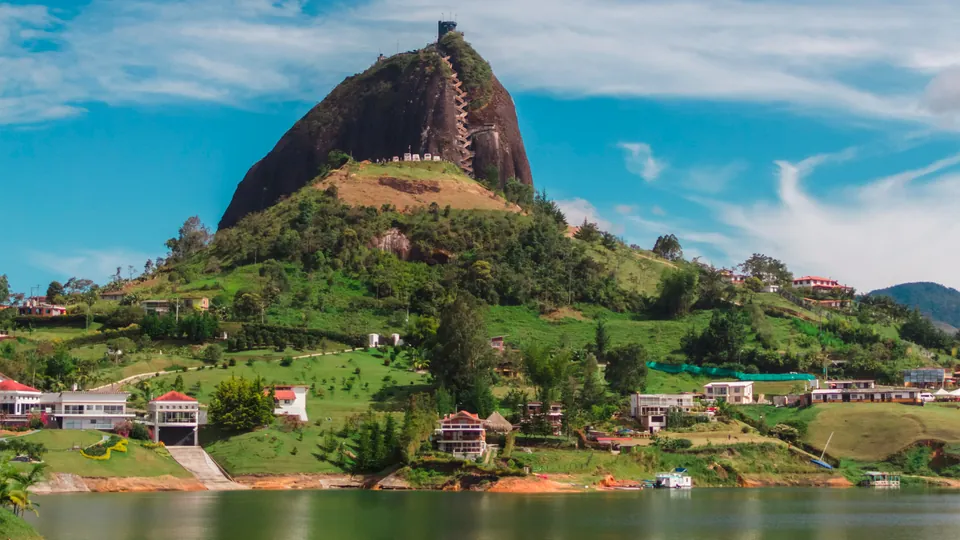 La Piedra del Peñol vista desde el embalse de Guatapé, con la escalera que sube por la grieta y las colinas verdes alrededor.